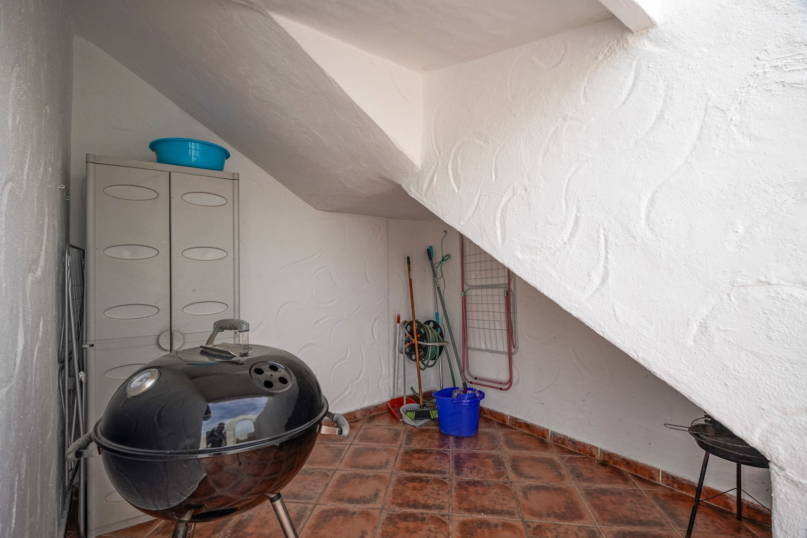 Zone de rangement extérieure sous l'escalier avec sol en terre cuite, armoire en résine et espace pour barbecue et matériel de nettoyage.