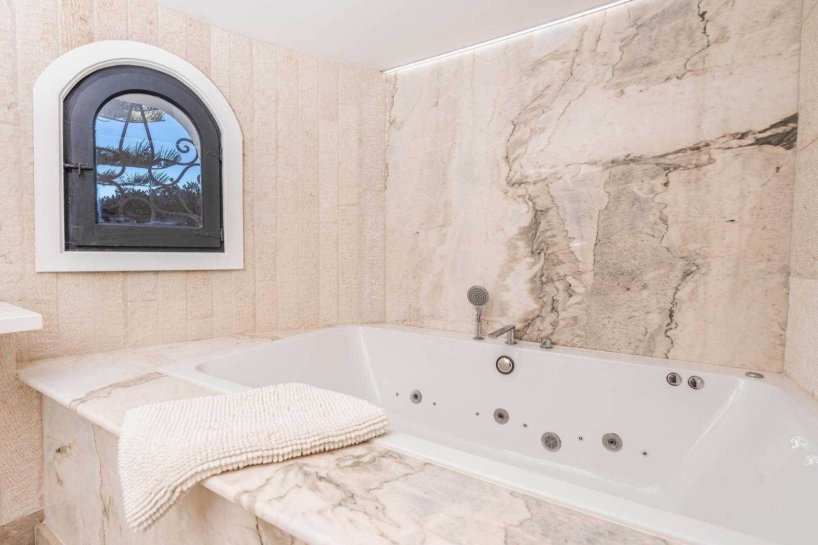 Baignoire d'hydromassage encastrée dans du marbre veiné, avec une fenêtre en arche traditionnelle et des murs en pierre naturelle éclairés par des LED.