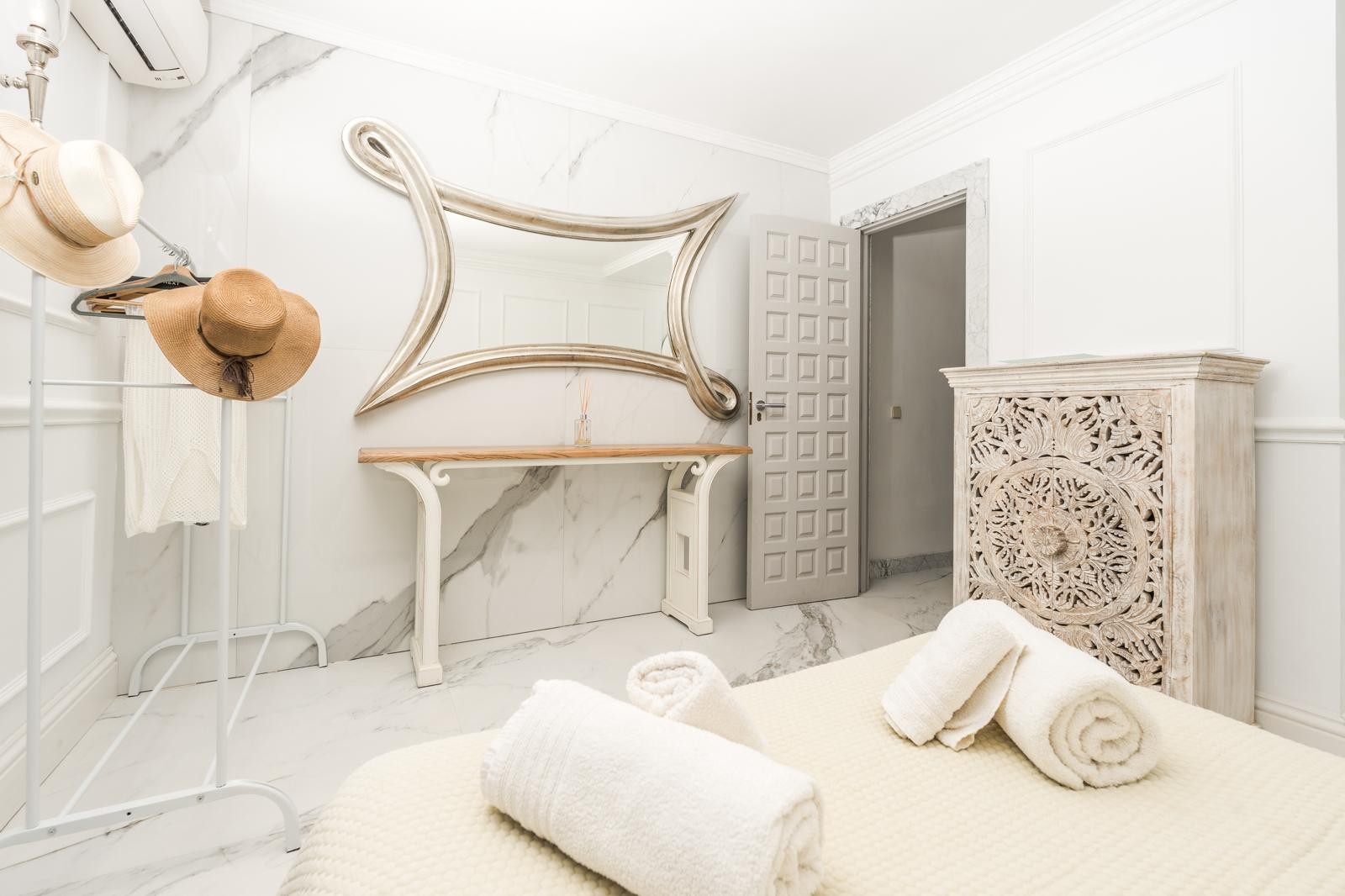 Chambre avec murs en marbre, miroir argenté aux formes organiques et commode en bois sculpté. Équipée de la climatisation et d'un sol élégant en grès cérame.