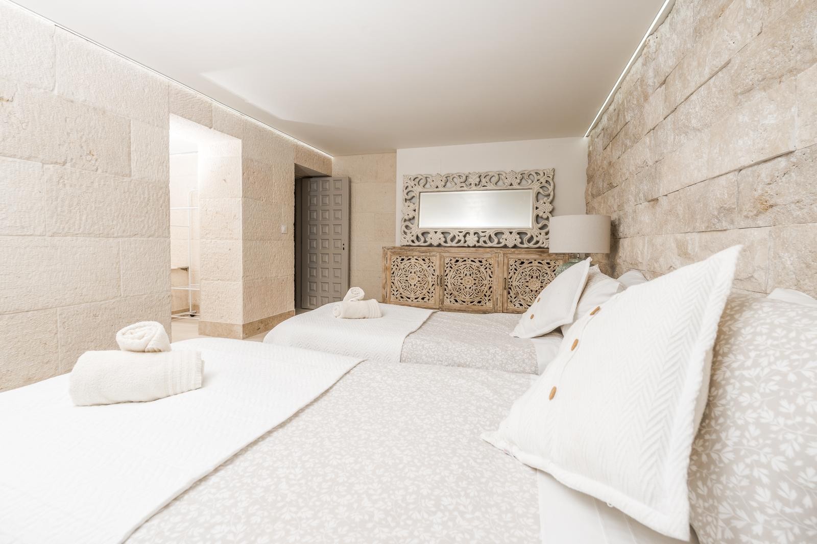 Chambre avec murs en pierre naturelle, lits jumeaux et buffet en bois sculpté. L'éclairage LED indirect souligne l'élégance des matériaux méditerranéens.