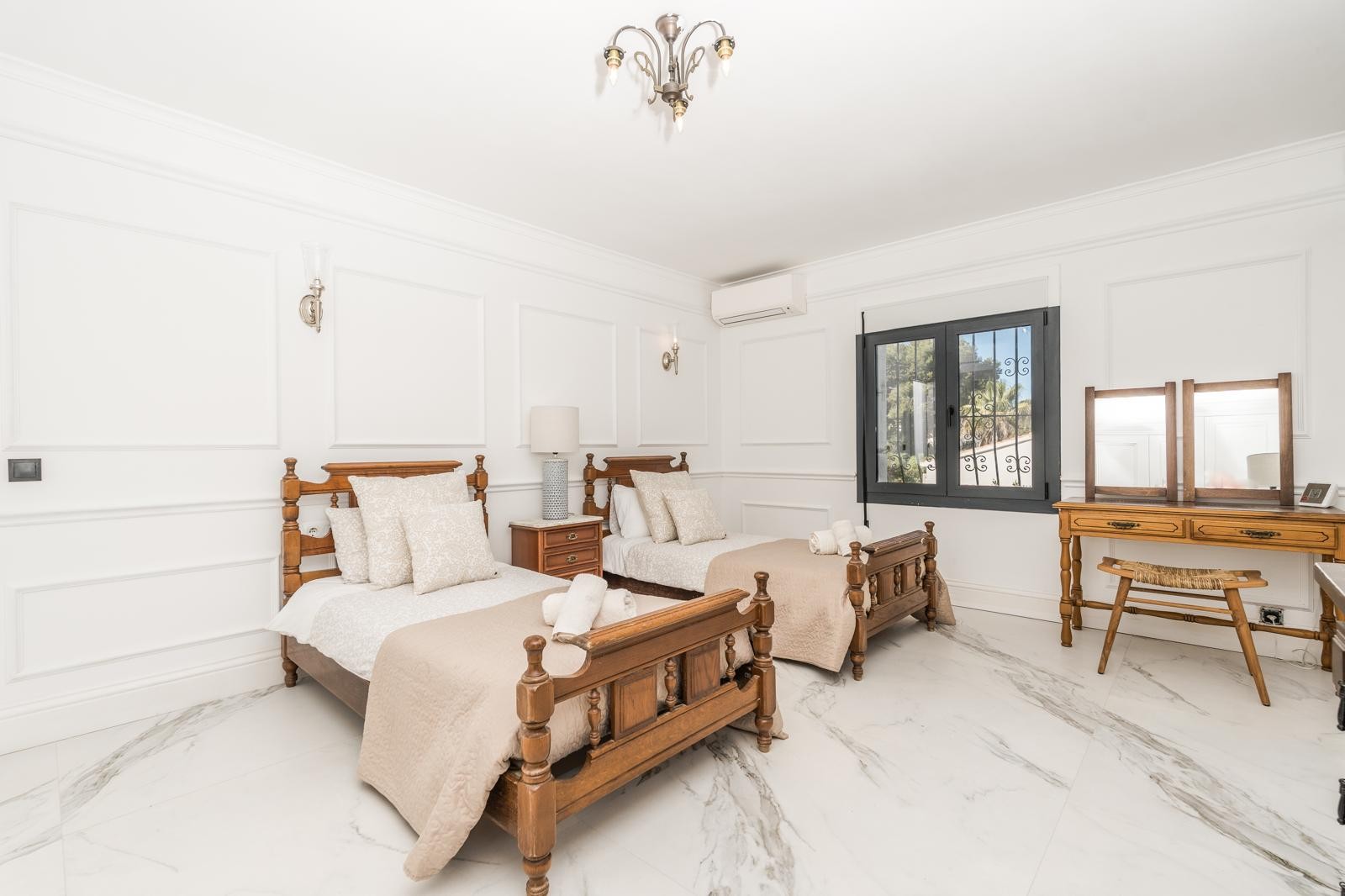 Chambre lumineuse avec deux lits simples en bois, sol en marbre veiné et murs ornés de moulures classiques. Équipée d'une coiffeuse en bois et de la climatisation individuelle.