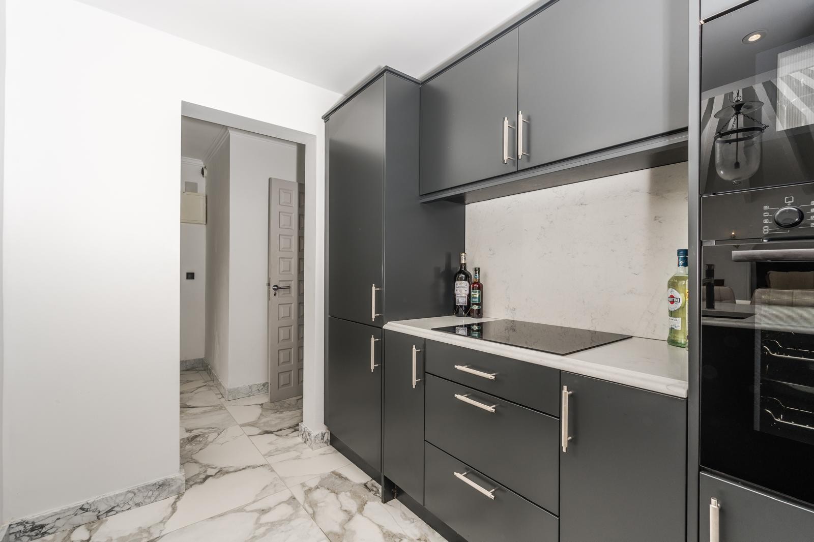 Cuisine contemporaine avec placards gris foncé, plan de travail en marbre blanc et plaque à induction sur un sol en marbre veiné.