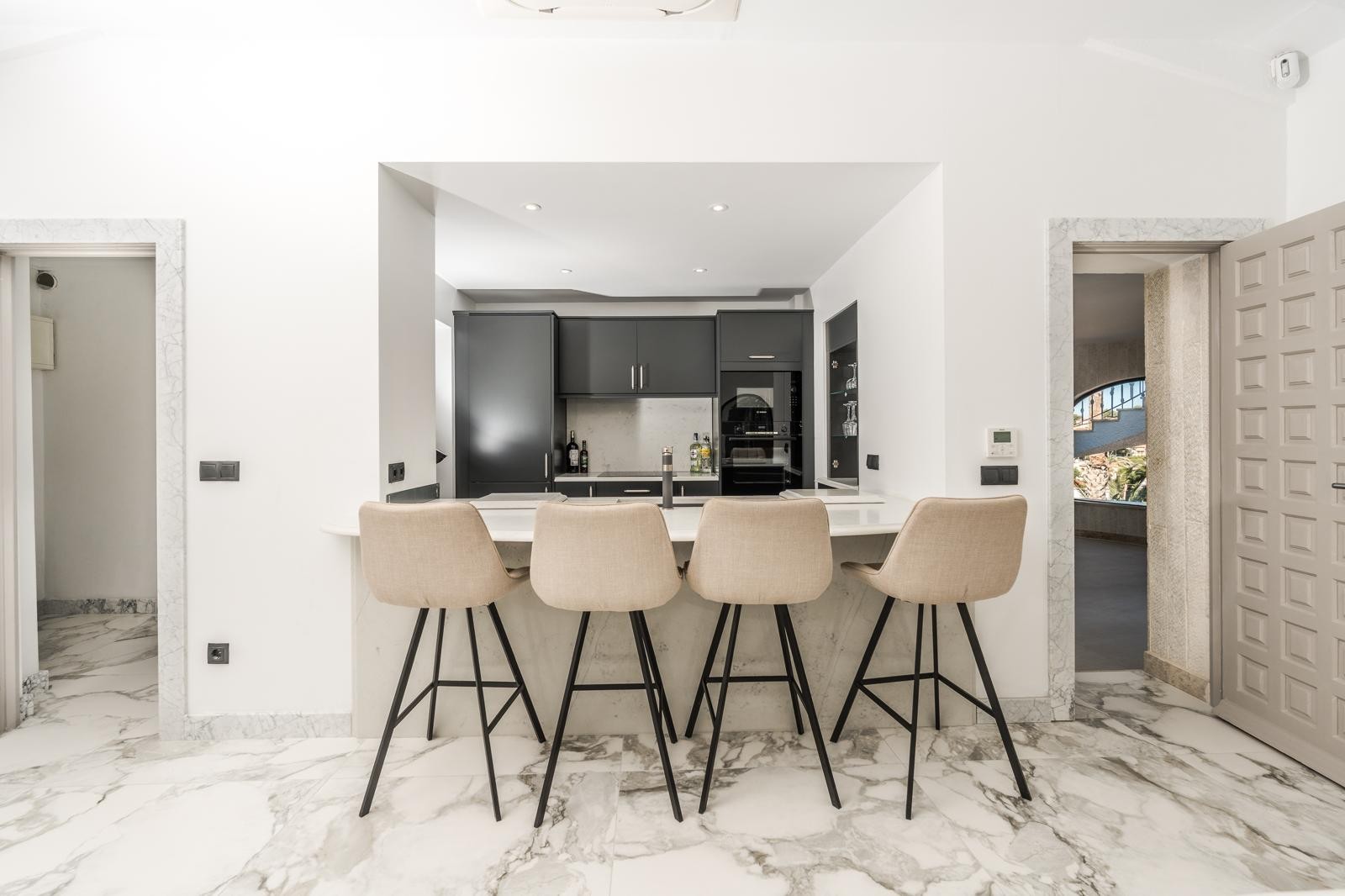 Cuisine semi-ouverte avec bar, tabourets rembourrés et plans de travail blancs. Le sol en marbre veiné et les encadrements de portes en pierre soulignent le luxe.