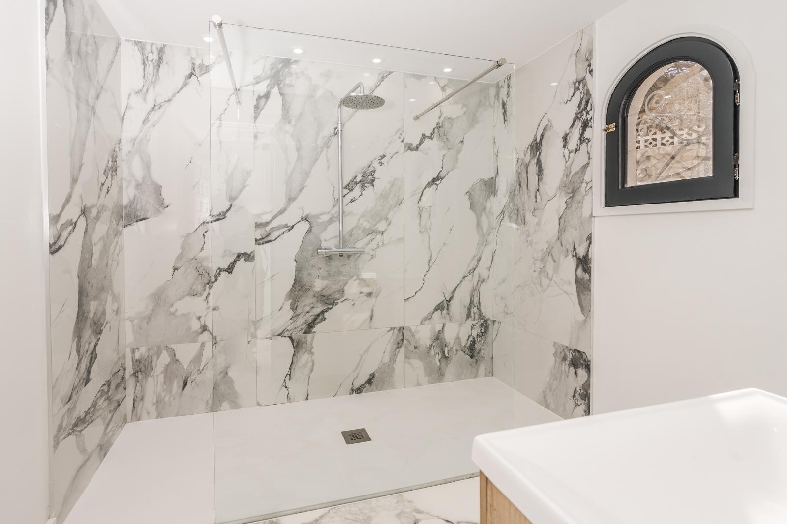 Douche à l'italienne avec parois en marbre grand format, paroi en verre fixe, robinetterie chromée et fenêtre cintrée au cadre noir.