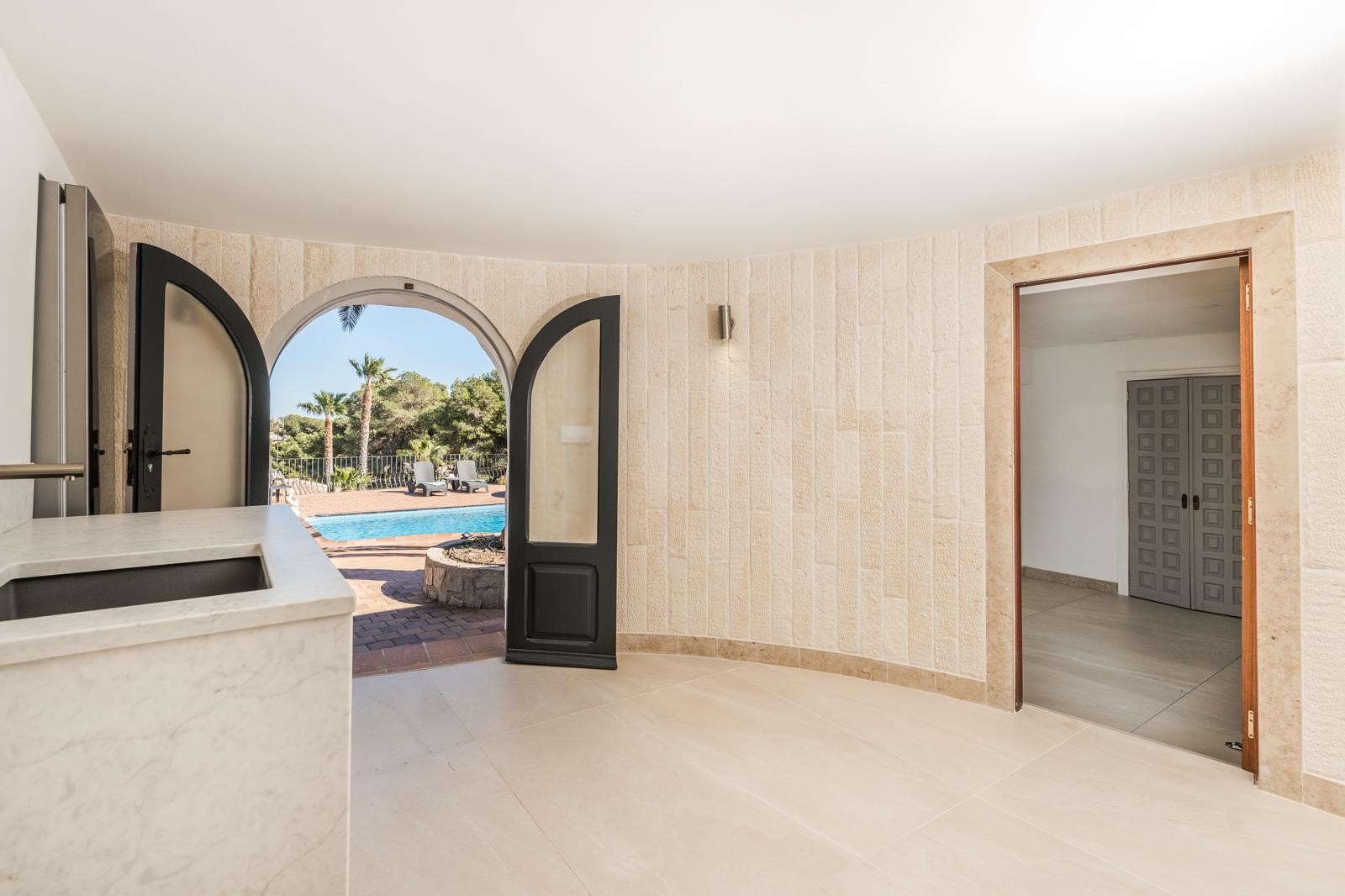 Espace intérieur avec murs en pierre naturelle et plan de travail en marbre, s'ouvrant sur la terrasse et la piscine par une porte cintrée.