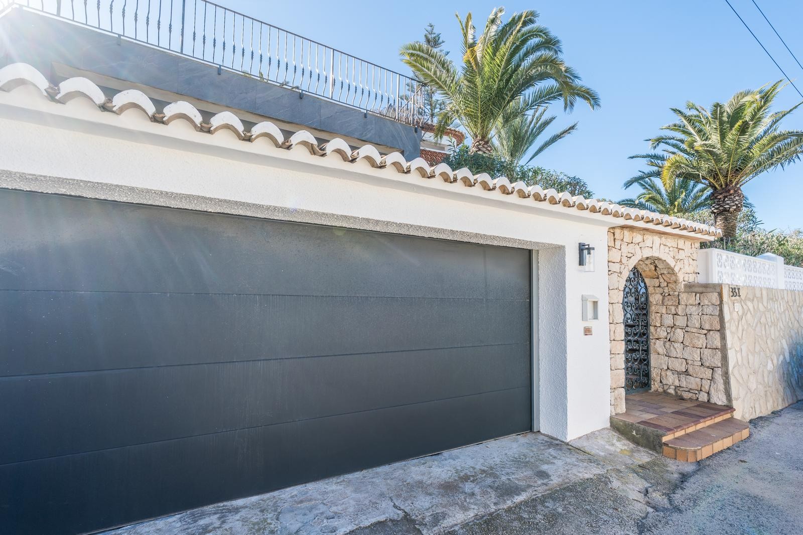 Façade extérieure avec porte de garage double gris anthracite, arche en pierre naturelle avec portillon en fer forgé et mur d'enceinte en pierre sous les palmiers.