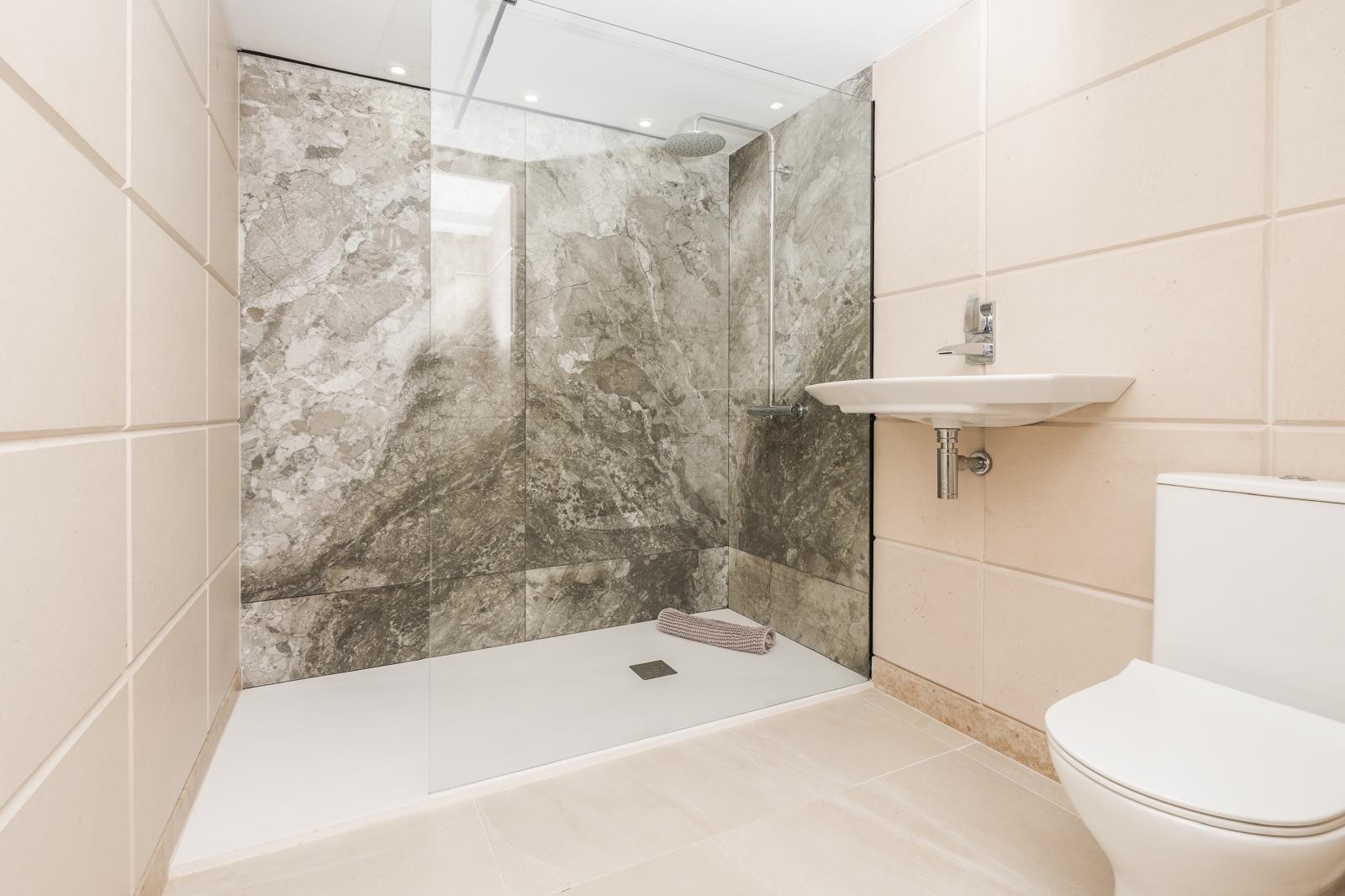 Salle de bain moderne avec douche à l'italienne, paroi en verre et carrelage effet marbre gris. Équipée d'un lavabo suspendu et d'un éclairage de plafond encastré.