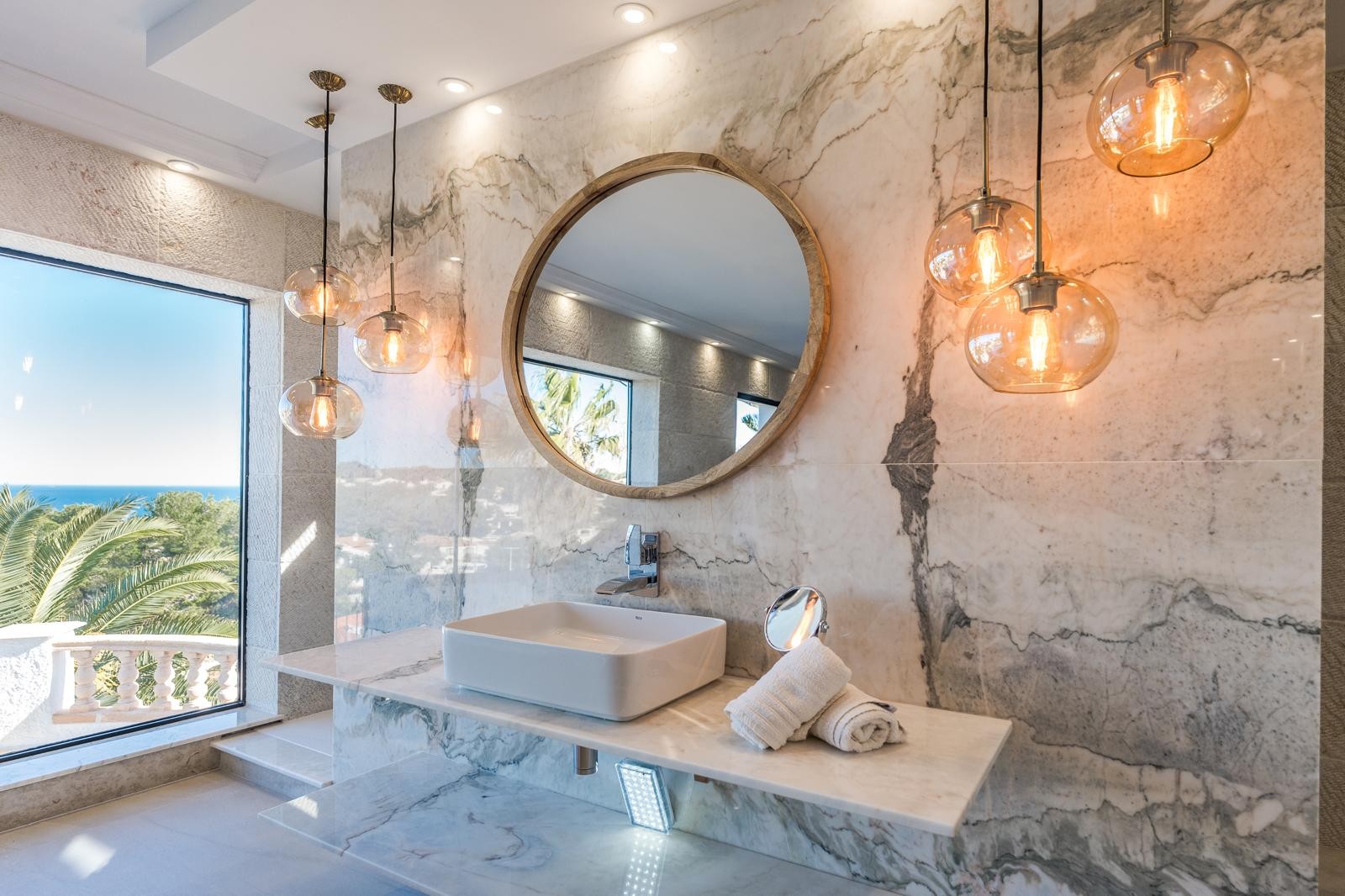 Salle de bains de design avec mur et plan de travail en marbre veiné, miroir circulaire en bois et suspensions en verre avec vue sur la mer.