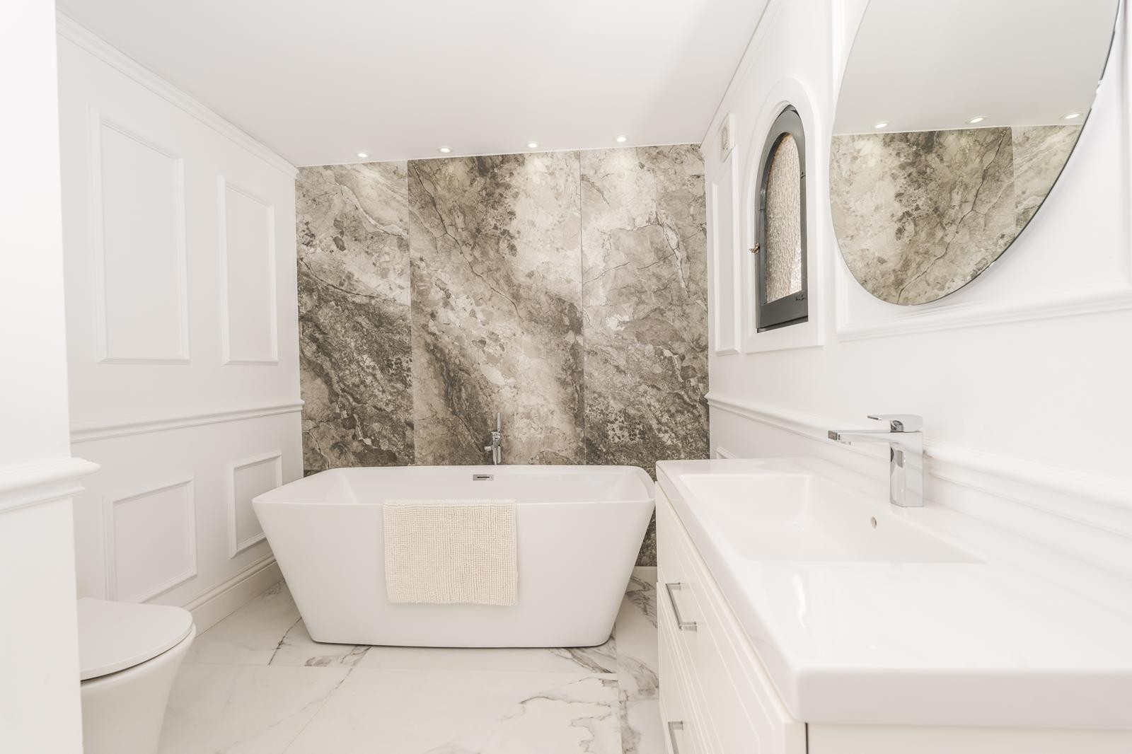 Salle de bains de la suite parentale avec baignoire îlot, mur en marbre veiné et moulures élégantes. Elle dispose d'un lavabo moderne, d'un miroir rond et d'une fenêtre en arche.