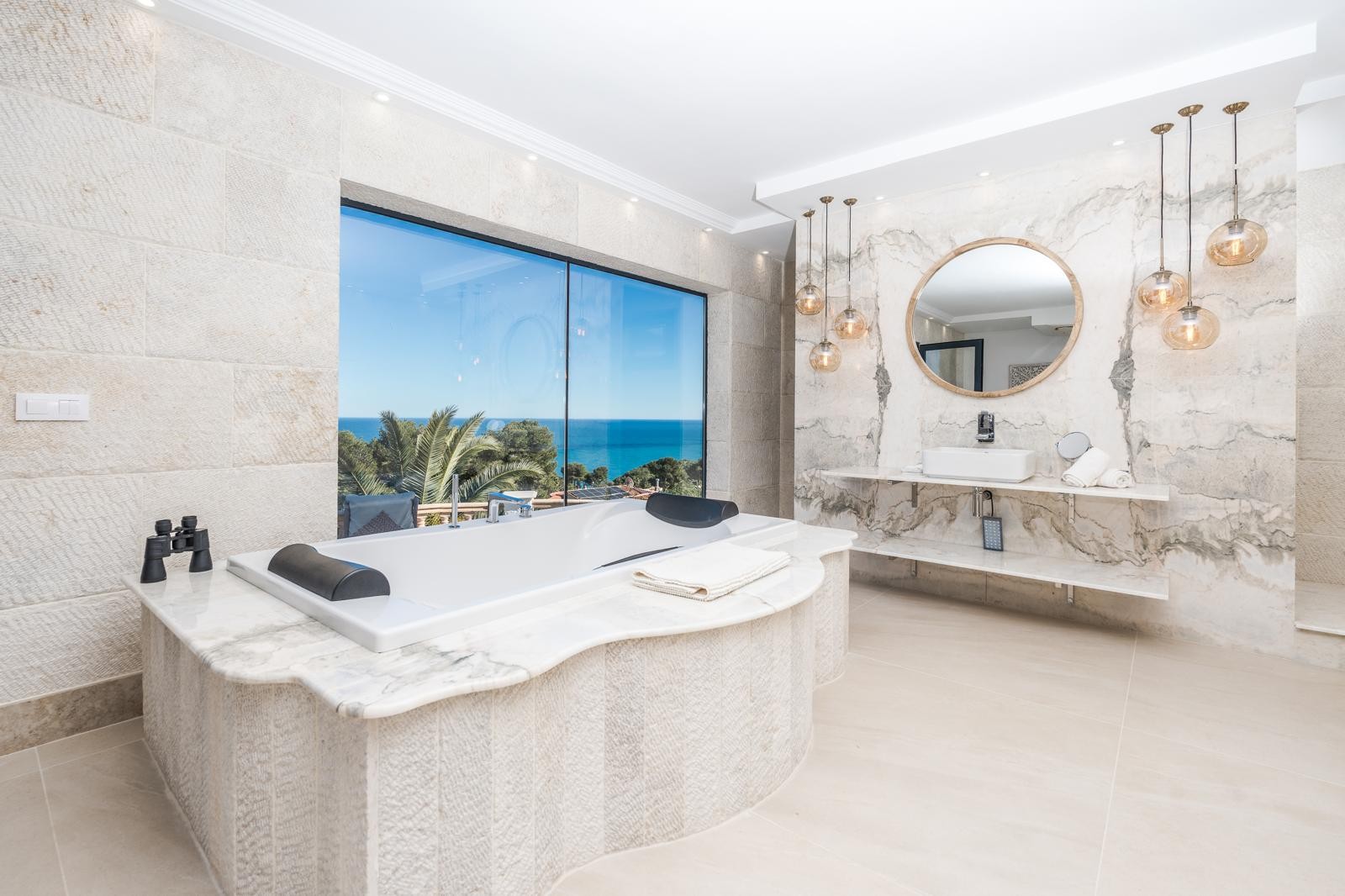 Salle de bains de la suite principale avec baignoire face à la mer, murs en marbre veiné, étagères en pierre et luminaires suspendus design.
