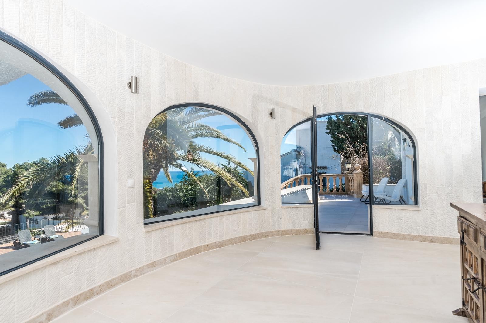 Salon circulaire aux murs en pierre naturelle, doté de grandes fenêtres en arche offrant une vue sur la mer et un accès à la terrasse ensoleillée.