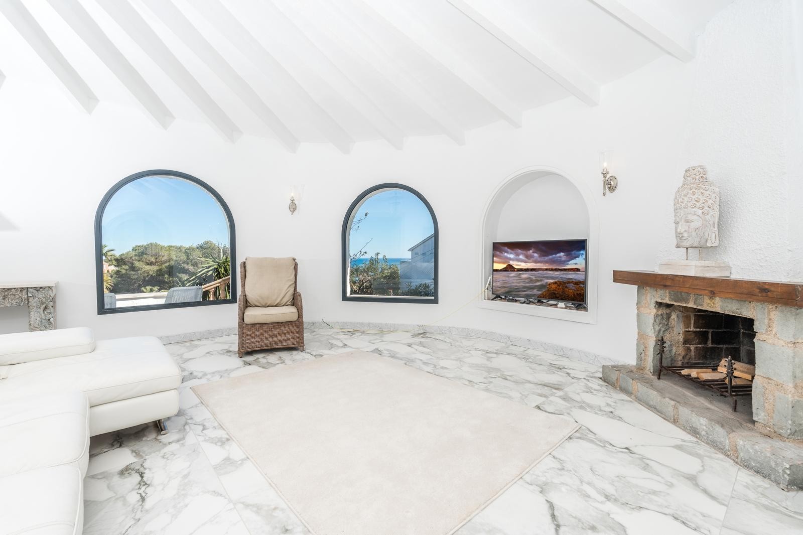 Salon circulaire avec plafond à poutres blanches, sol en marbre et cheminée en pierre. Des fenêtres cintrées offrent une vue dégagée et une luminosité naturelle abondante.