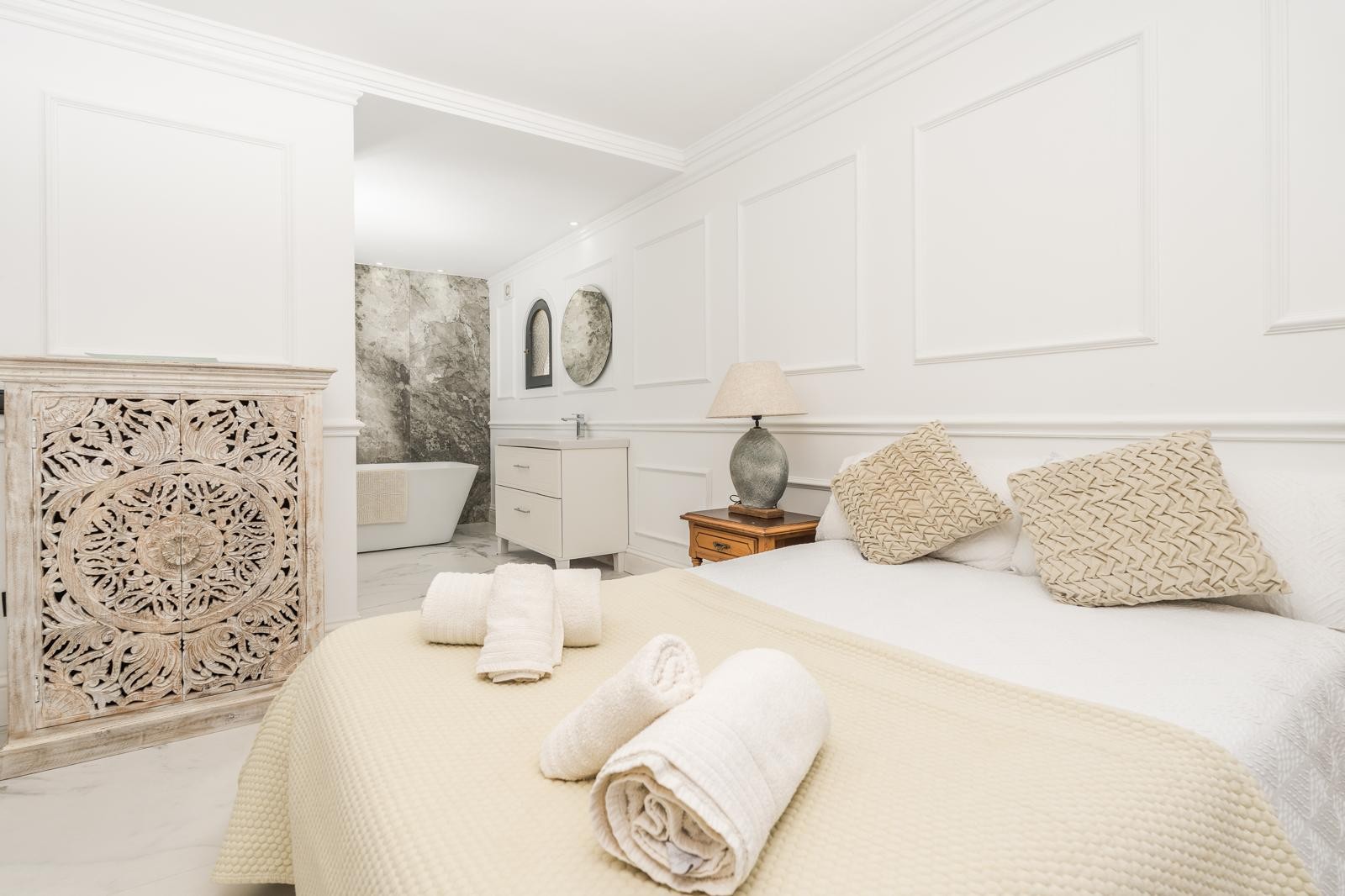 Suite parentale avec salle de bain ouverte, comprenant une baignoire îlot devant un mur en marbre gris et des murs ornés de moulures élégantes et d'un meuble sculpté.