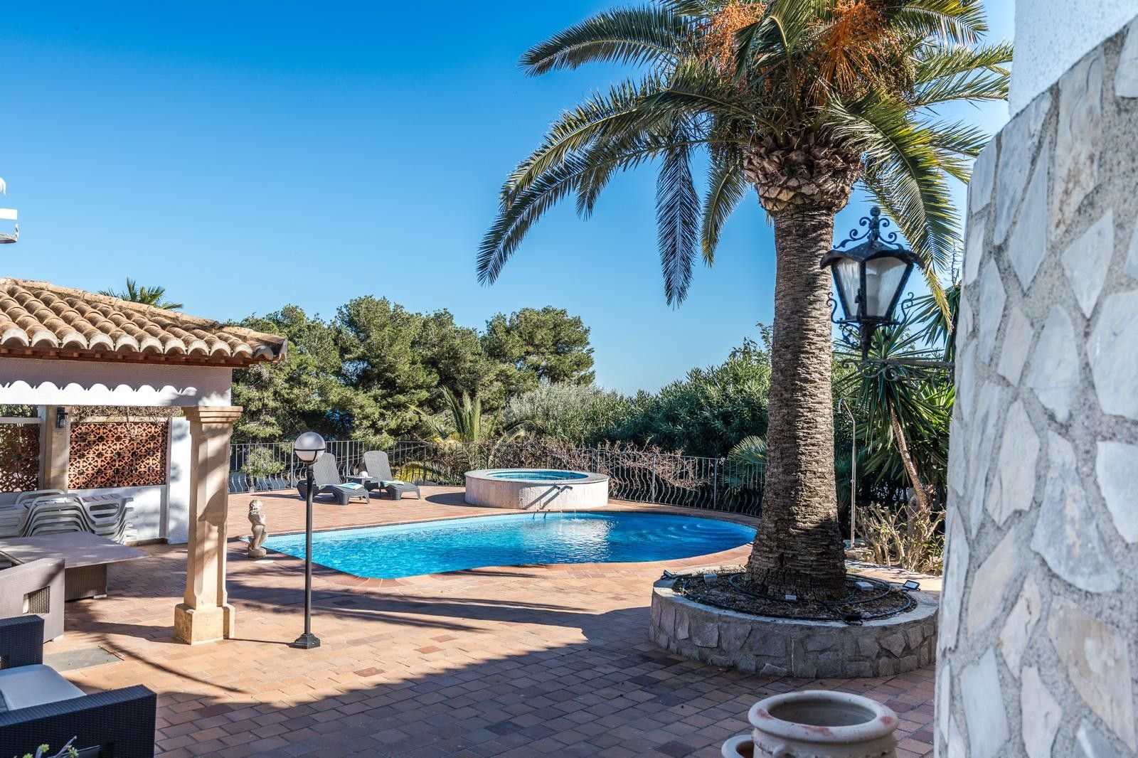 Terrasse extérieure avec piscine, jacuzzi circulaire et porche couvert, nichée dans un jardin méditerranéen avec palmiers et dallage en terre cuite.