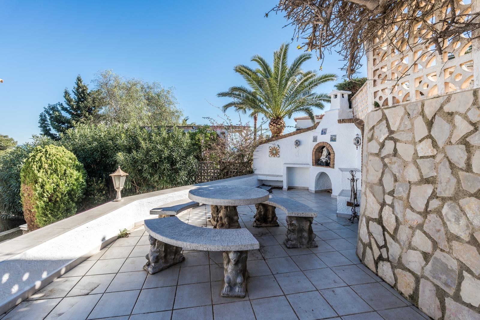 Terrasse extérieure avec un ensemble table et bancs en pierre sculptée, un barbecue maçonné intégré et des murs en pierre naturelle dans un cadre méditerranéen.