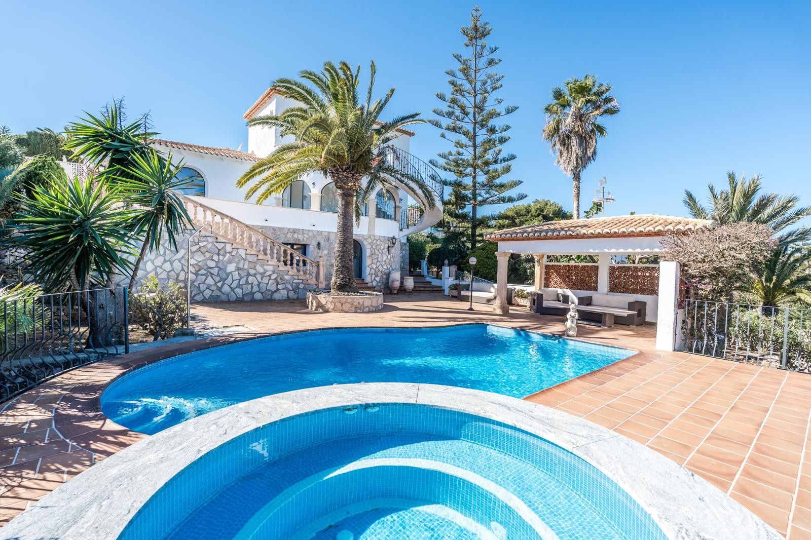 Vue extérieure de la piscine rénovée et du jacuzzi circulaire, intégrés dans une terrasse en terre cuite avec salon couvert et végétation méditerranéenne.