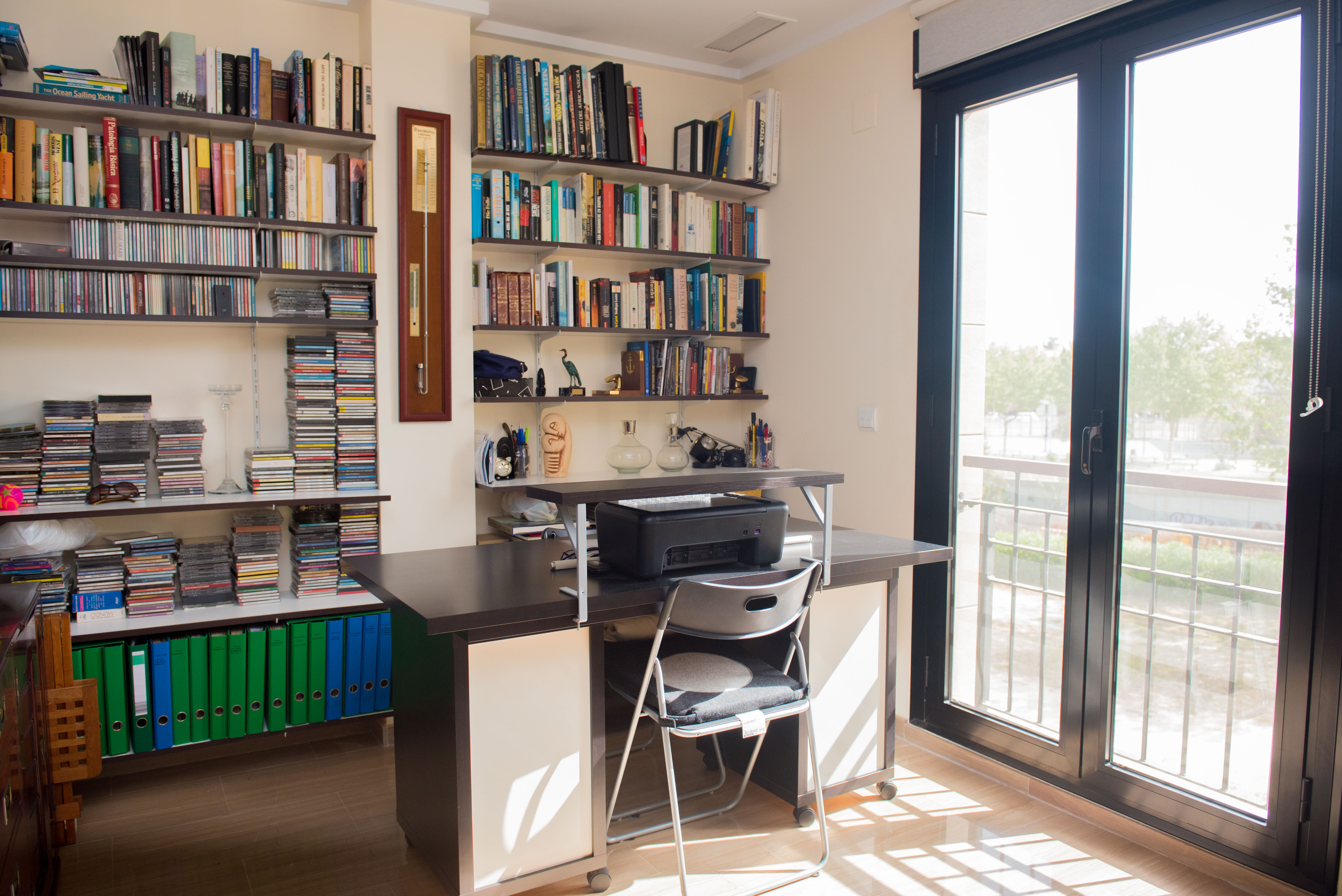 Bureau lumineux avec un grand bureau en bois foncé, des bibliothèques murales remplies de livres et de CD, et une porte vitrée menant à un balcon extérieur. Bureau lumineux avec un grand bureau en bois foncé, des bibliothèques murales remplies de livres et de CD, et une porte vitrée menant à un balcon extérieur.