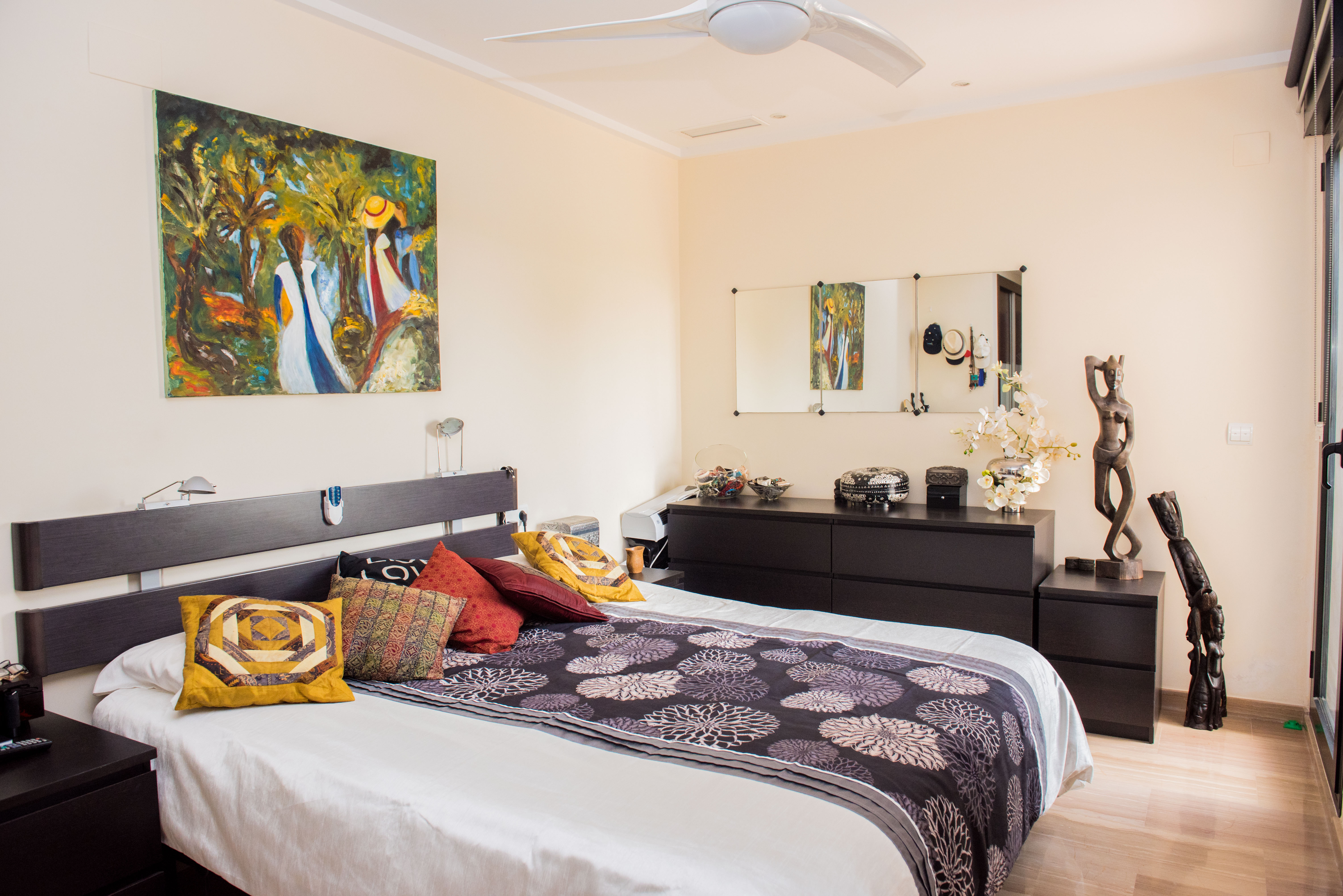 Chambre avec lit double, tête de lit en bois sombre et ventilateur de plafond. Comprend une commode assortie, une peinture murale colorée et un sol clair.