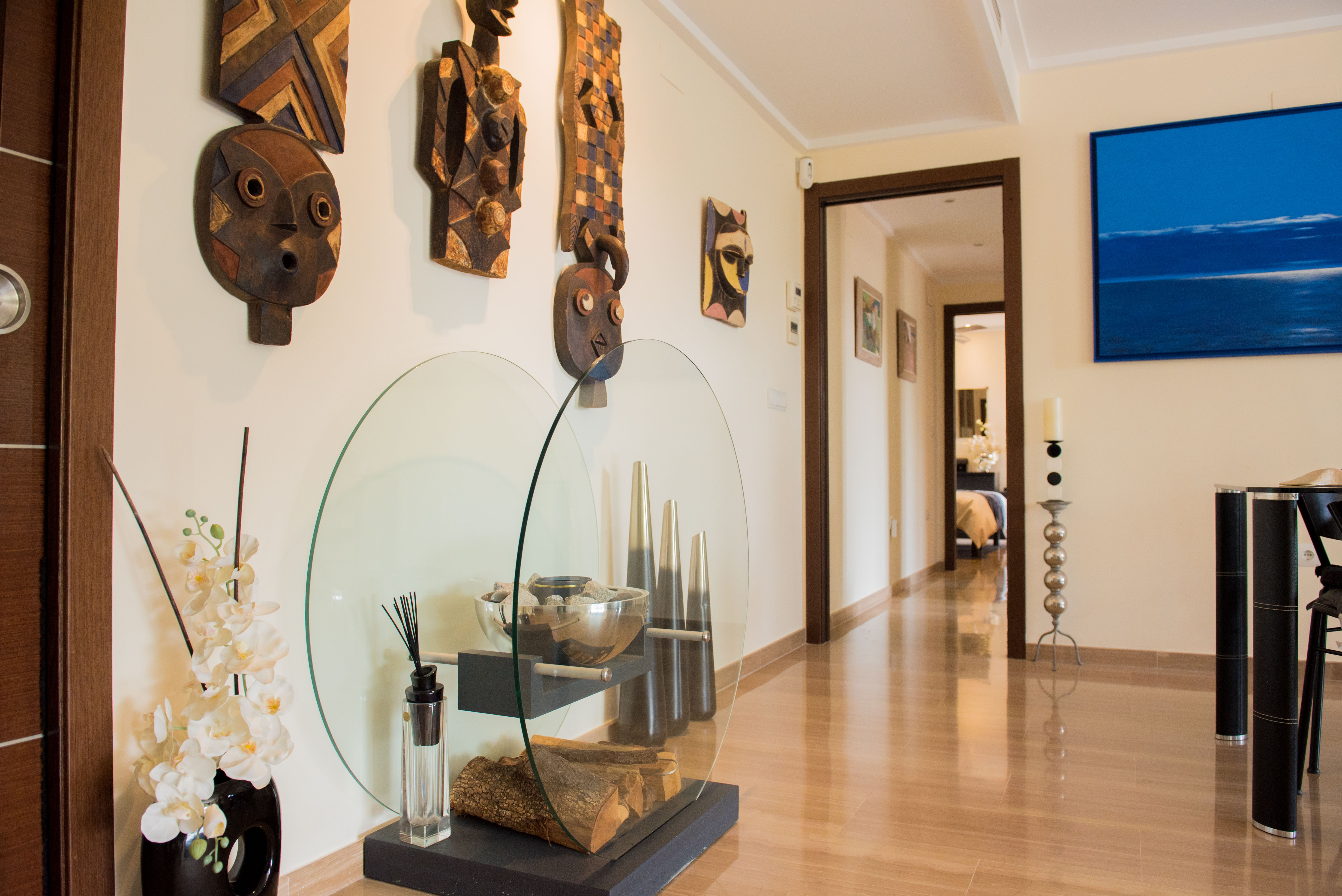 Couloir lumineux avec carrelage brillant effet bois, orné de masques tribaux en bois et d'une vitrine en verre contemporaine avec bûches et orchidées. Une œuvre abstraite bleue est visible. Couloir lumineux avec carrelage brillant effet bois, orné de masques tribaux en bois et d'une vitrine en verre contemporaine avec bûches et orchidées. Une œuvre abstraite bleue est visible.