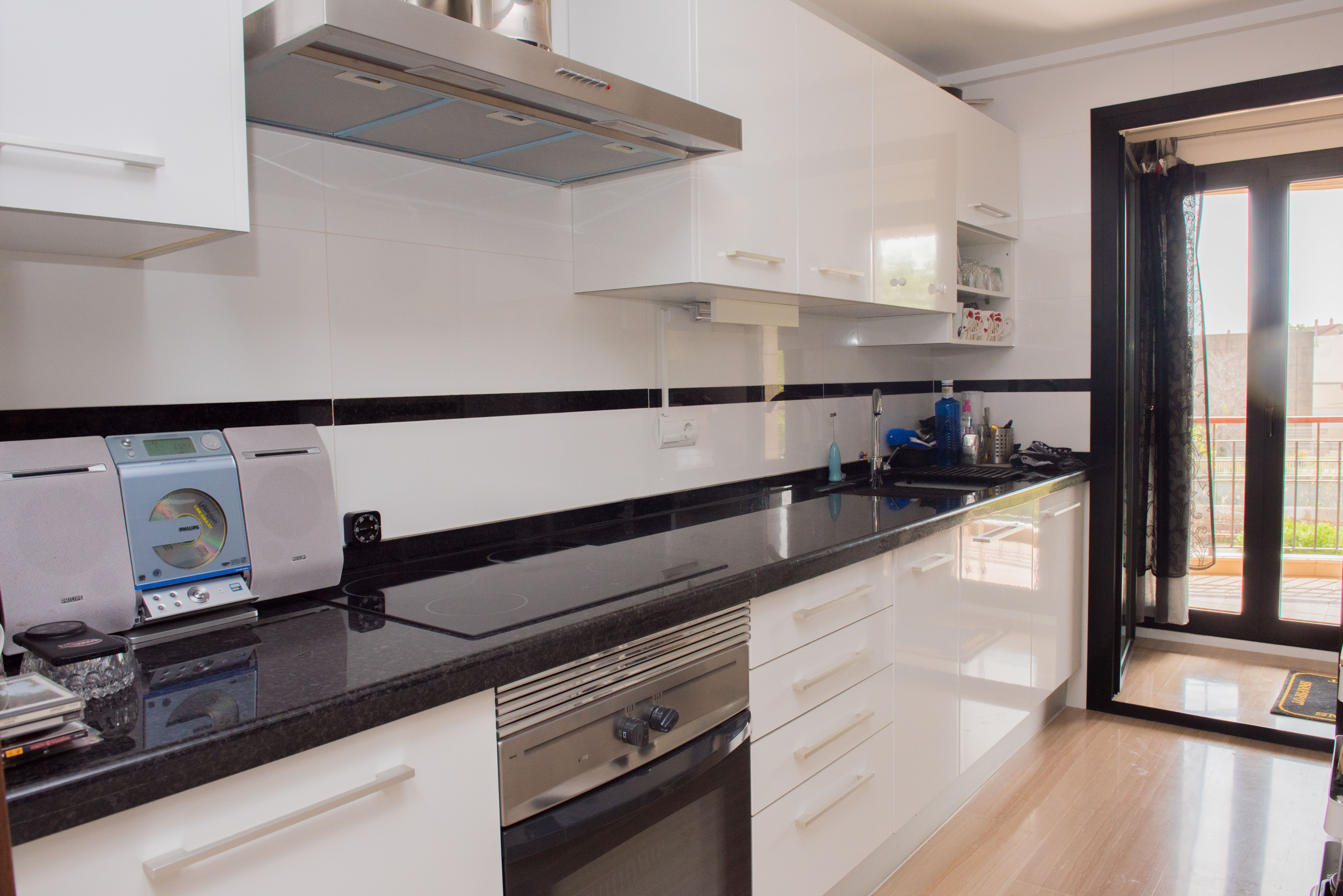 Cuisine moderne avec meubles blancs brillants, plan de travail en granit noir et électroménager en inox. Accès direct à un balcon par une porte vitrée.