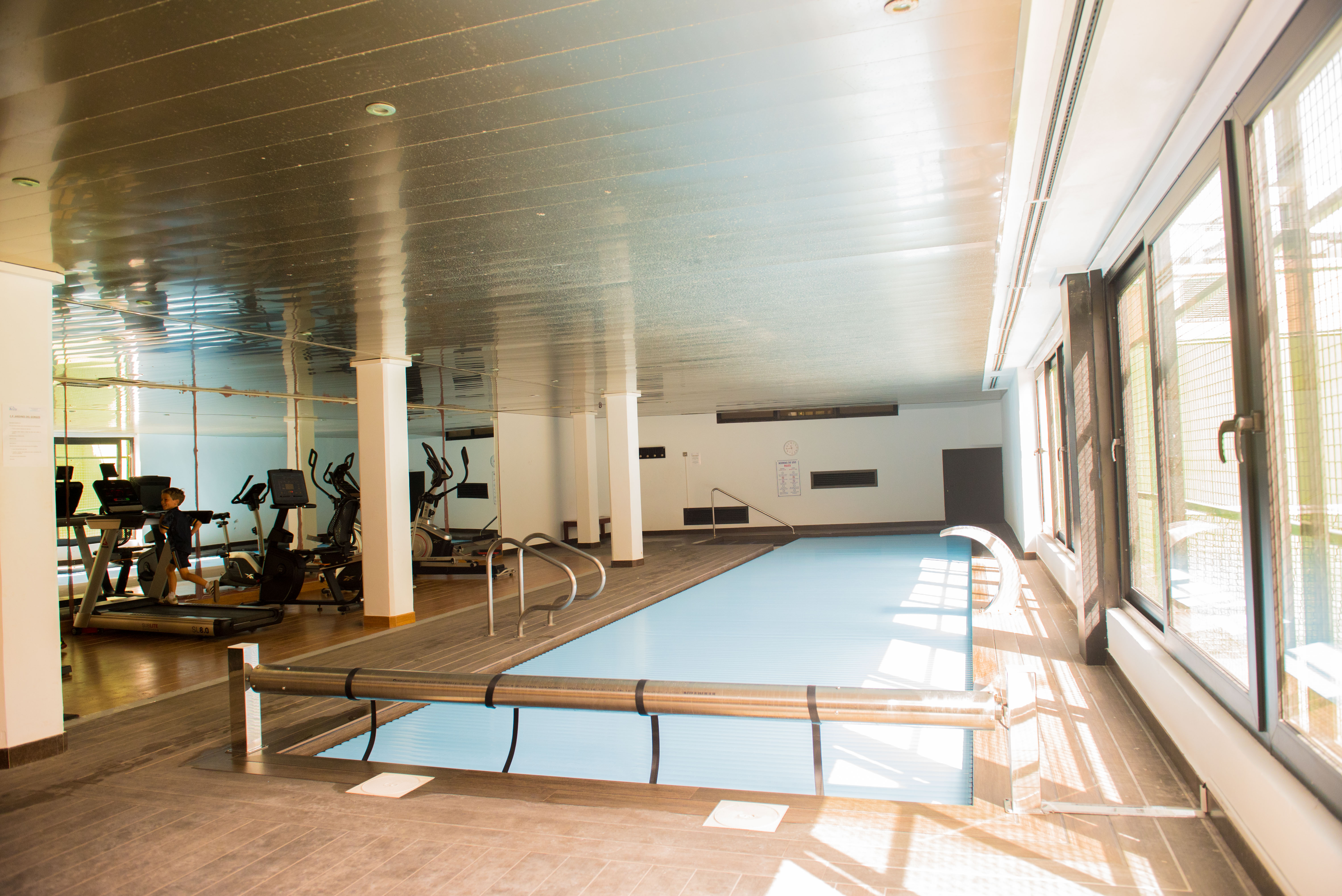 Espace bien-être intérieur avec piscine chauffée couverte et salle de sport équipée de machines de cardio, sous un plafond métallique réfléchissant.