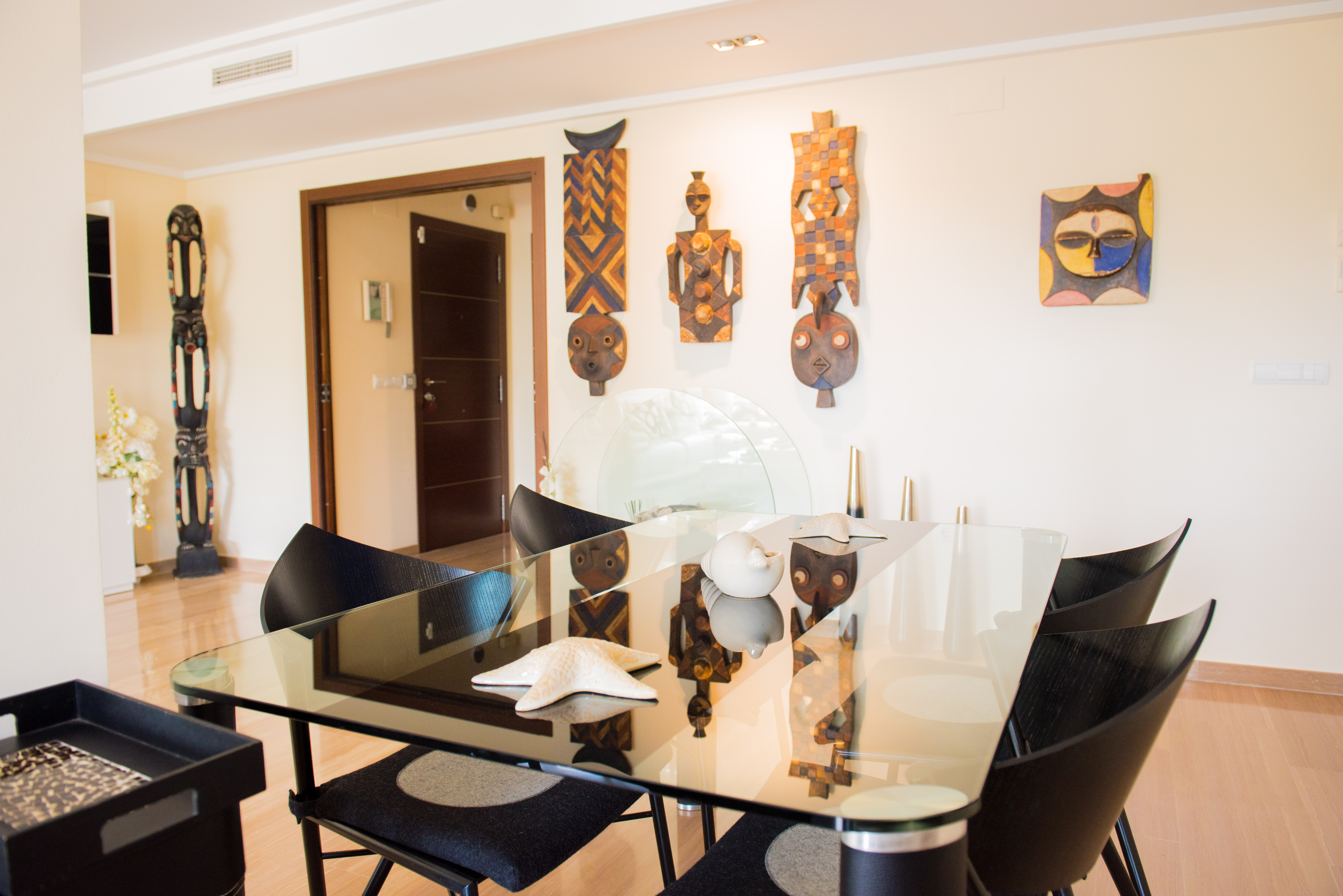 Espace salle à manger contemporain avec table en verre, chaises noires élégantes et parquet clair. Des masques ethniques décorent le mur. Espace salle à manger contemporain avec table en verre, chaises noires élégantes et parquet clair. Des masques ethniques décorent le mur.