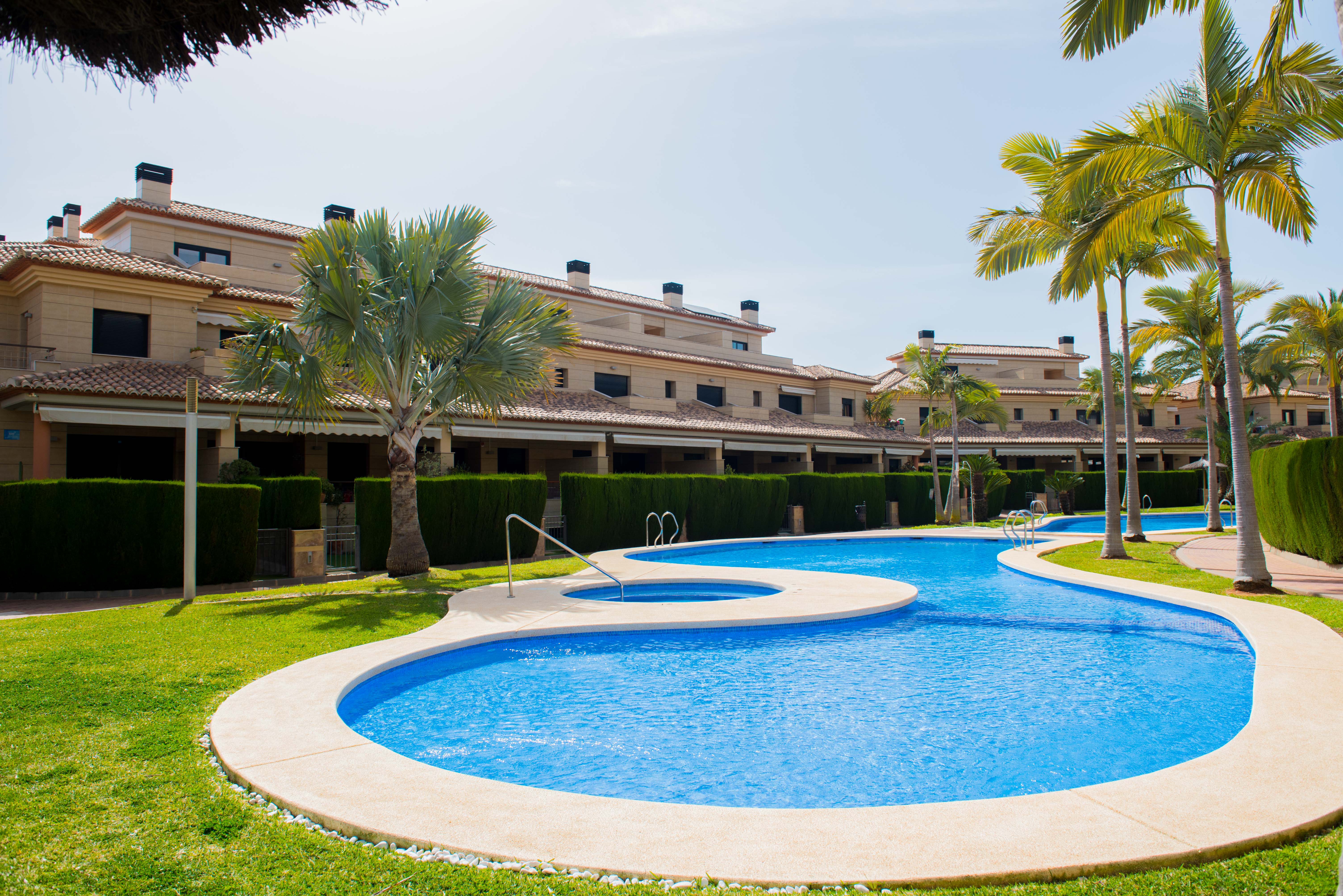 Grande piscine communautaire avec spa intégré, entourée de pelouses et de palmiers, située devant des résidences aux façades de pierre naturelle.