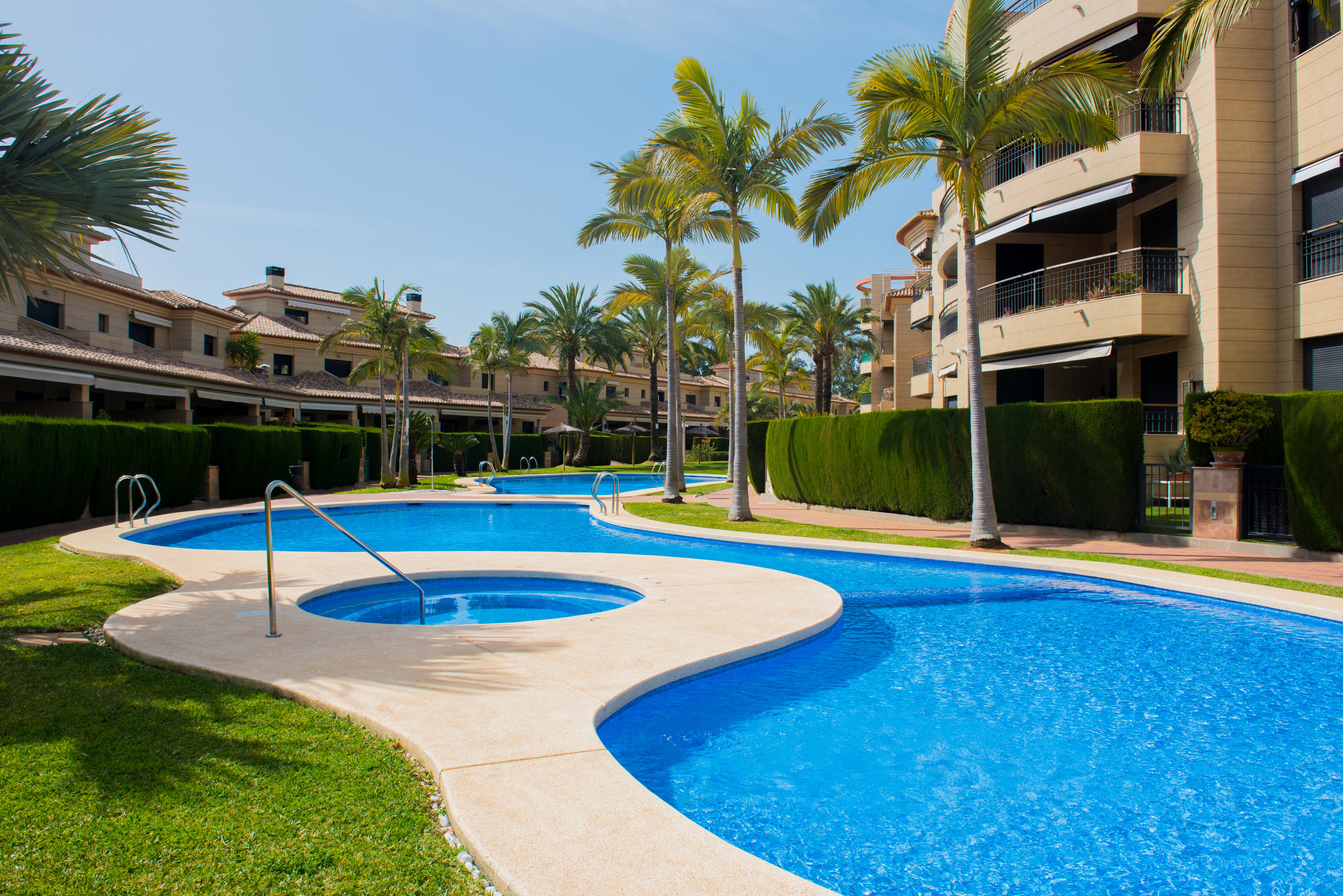 Piscine communautaire aux formes courbes avec jacuzzi intégré, entourée de jardins paysagers, de palmiers et de résidences aux façades en pierre.