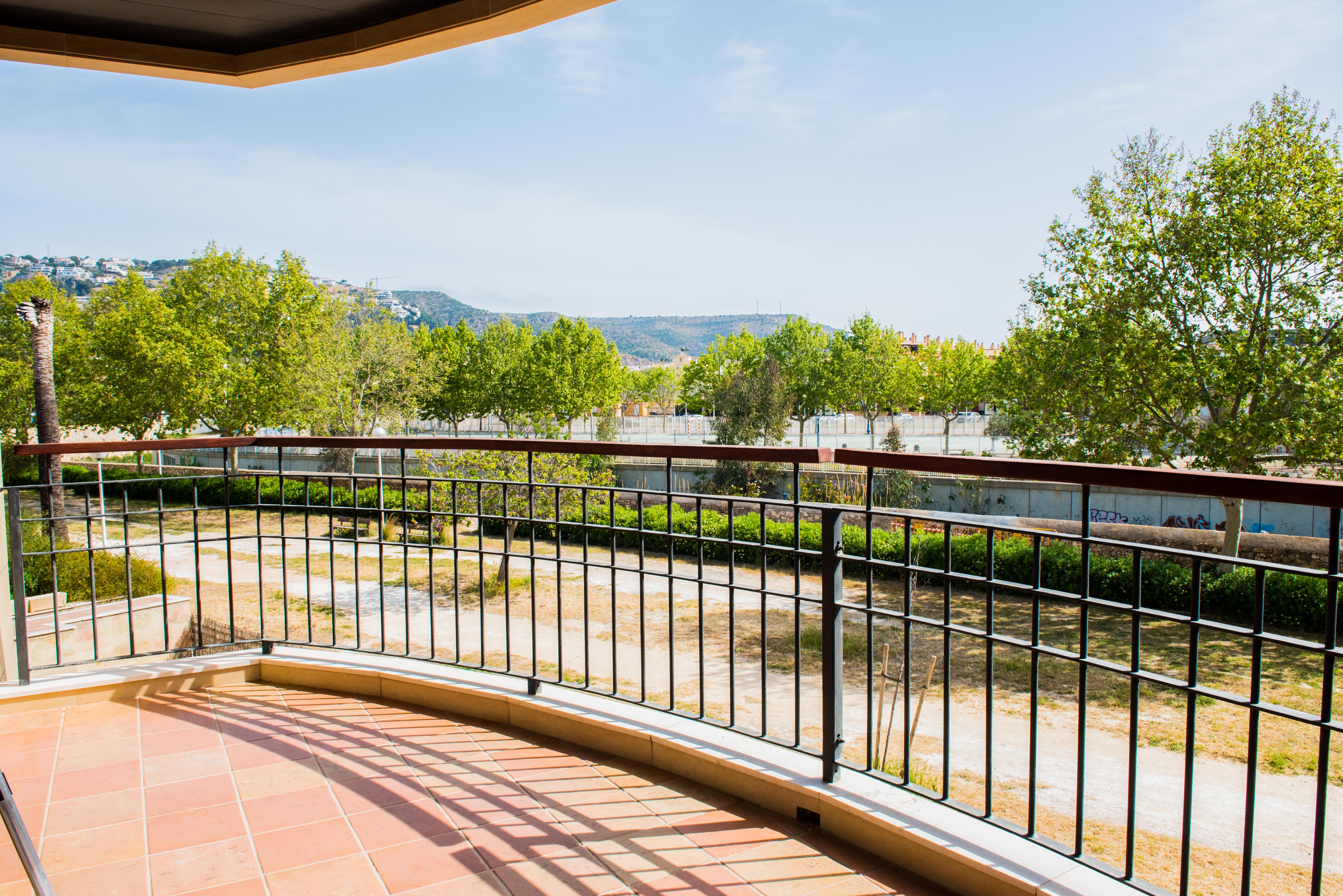 Terrasse couverte avec sol en terre cuite et garde-corps en fer forgé avec main courante en bois, offrant une vue sur les espaces verts et les montagnes.
