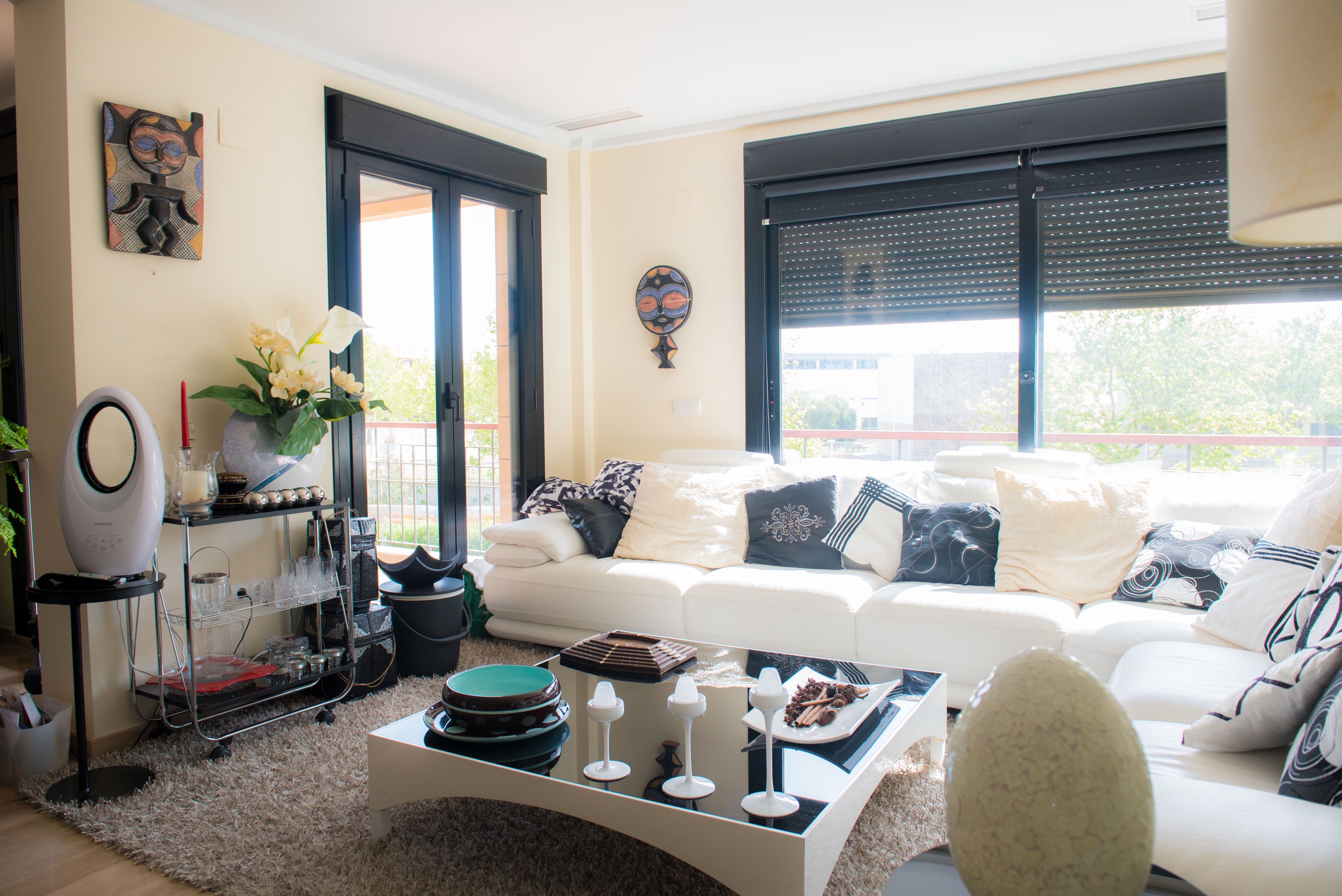 Vaste salon avec canapé blanc et coussins variés, tapis moelleux et table basse moderne. Grandes fenêtres à cadres noirs donnant sur la verdure extérieure. Vaste salon avec canapé blanc et coussins variés, tapis moelleux et table basse moderne. Grandes fenêtres à cadres noirs donnant sur la verdure extérieure.