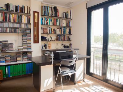 Bureau lumineux avec un grand bureau en bois foncé, des bibliothèques murales remplies de livres et de CD, et une porte vitrée menant à un balcon extérieur.