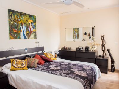 Chambre principale avec lit double, tête de lit foncée et tables de chevet assorties. Un tableau abstrait décore le mur, avec commode, miroirs et ventilateur de plafond. Revêtement de sol imitation bois clair.