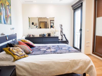 Chambre principale avec lit double, tête de lit sombre à lampes intégrées, et accès direct à une terrasse. Inclut une armoire encastrée bois/verre et un sol stratifié clair.