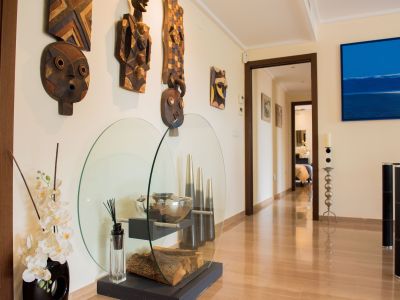 Couloir lumineux avec carrelage brillant effet bois, orné de masques tribaux en bois et d'une vitrine en verre contemporaine avec bûches et orchidées. Une œuvre abstraite bleue est visible.