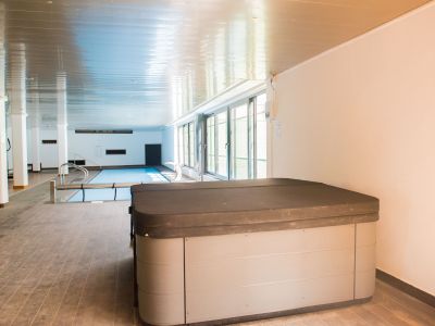Jacuzzi couvert au premier plan, avec piscine intérieure chauffée et grandes fenêtres apportant lumière naturelle à cet espace bien-être.