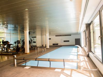 Piscine intérieure couverte, avec deck en bois. De larges baies vitrées inondent l'espace de lumière naturelle, et une salle de sport se reflète dans le miroir en arrière-plan.