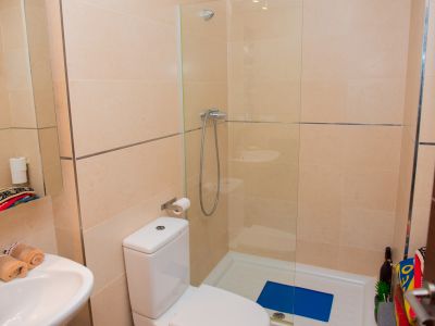 Salle de bain moderne avec carrelage mural beige clair et accents métalliques. Comprend des toilettes blanches, un lavabo avec robinet chromé et une douche vitrée avec receveur blanc.