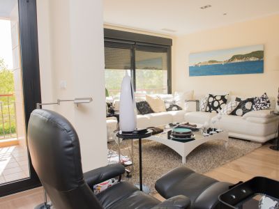 Séjour contemporain avec canapé d'angle blanc, table basse en verre noir et sol carrelé imitation bois clair. Accès direct à une terrasse avec balustrade métallique et vue sur la végétation.