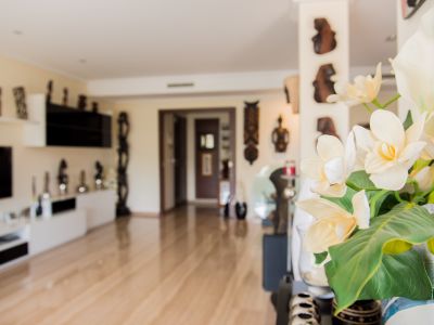 Vase au premier plan avec d'élégantes fleurs artificielles blanches et jaunes. Intérieur lumineux avec parquet clair, menant à un salon moderne agrémenté d'art mural décoratif.