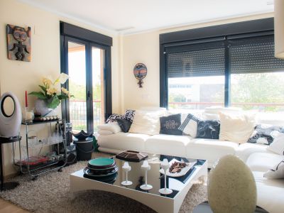 Vaste salon avec canapé blanc et coussins variés, tapis moelleux et table basse moderne. Grandes fenêtres à cadres noirs donnant sur la verdure extérieure.