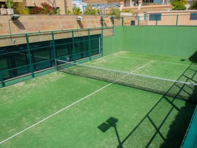 Vue de la piste de padel extérieure de la communauté, avec son gazon artificiel vert vif, son filet et ses murs en maille verte robuste, des bâtiments résidentiels étant visibles à l'arrière-plan ensoleillé.