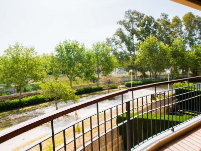 Vue depuis une terrasse privée sur de luxuriants jardins communautaires avec des arbres matures, un sentier sinueux et un mur en pierre, sous un ciel lumineux. La balustrade est métallique et le sol carrelé.