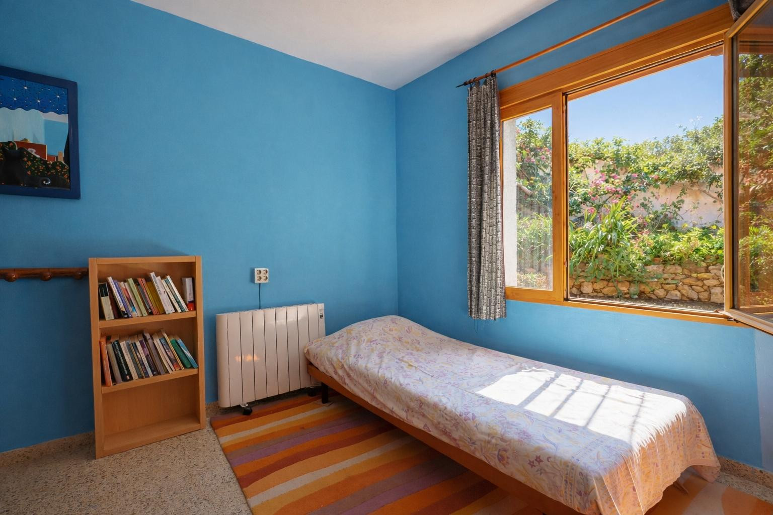 Chambre aux murs bleu vif avec lit simple et étagère en bois. La fenêtre à cadre en bois s'ouvre sur un jardin méditerranéen avec des murets en pierre.