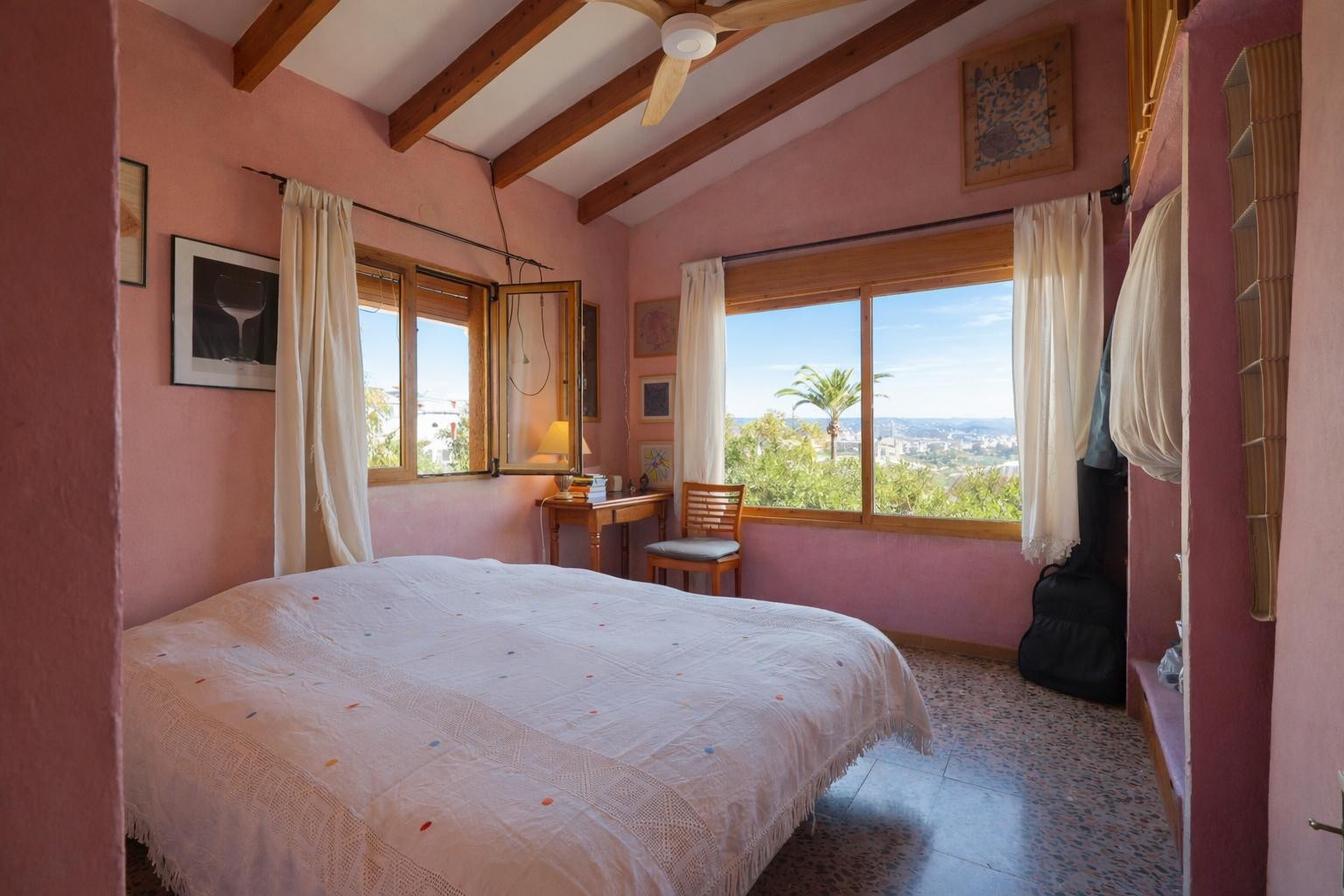 Chambre avec poutres en bois apparentes, murs aux tons chauds et grandes fenêtres offrant une vue dégagée sur le paysage méditerranéen.