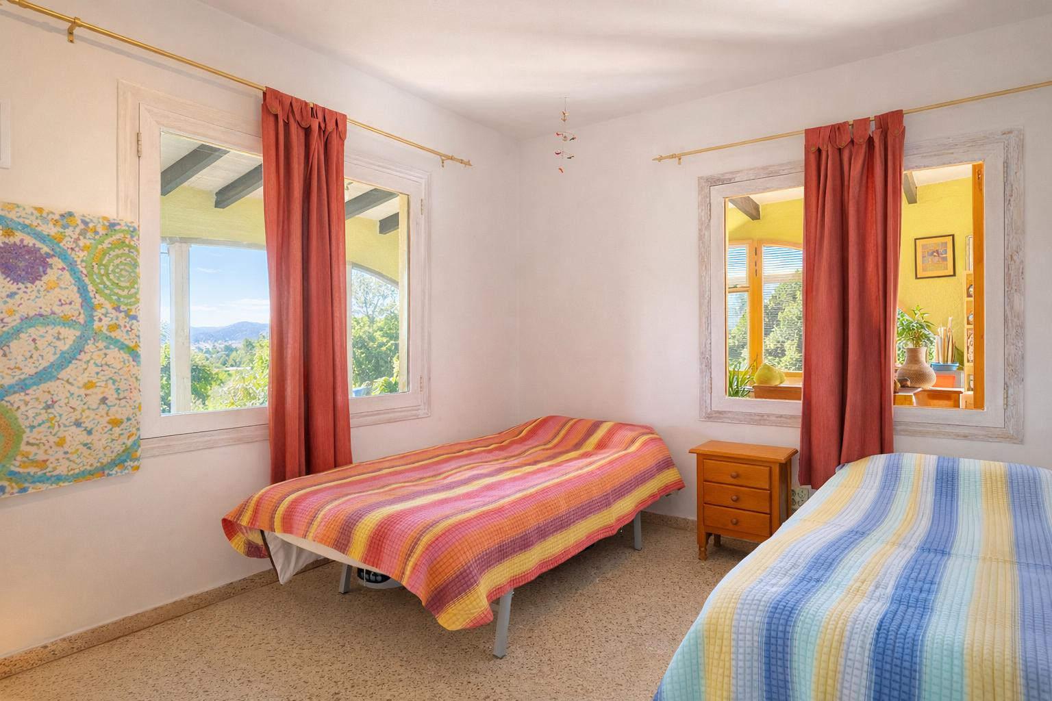 Chambre lumineuse avec deux lits simples, une table de chevet en bois et des fenêtres aux cadres blancs offrant une vue sur le paysage et l'intérieur.