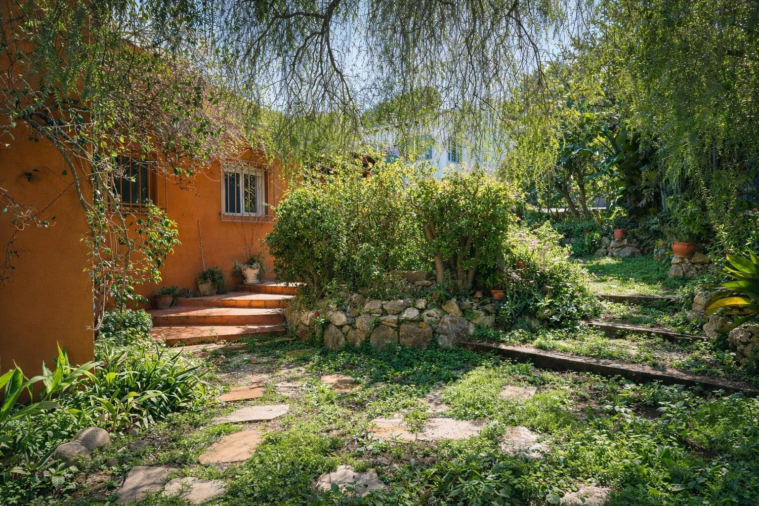 Jardin méditerranéen avec sentiers en pierre et marches en bois, entouré d'une végétation dense et de murets en pierre naturelle près de la façade ocre.