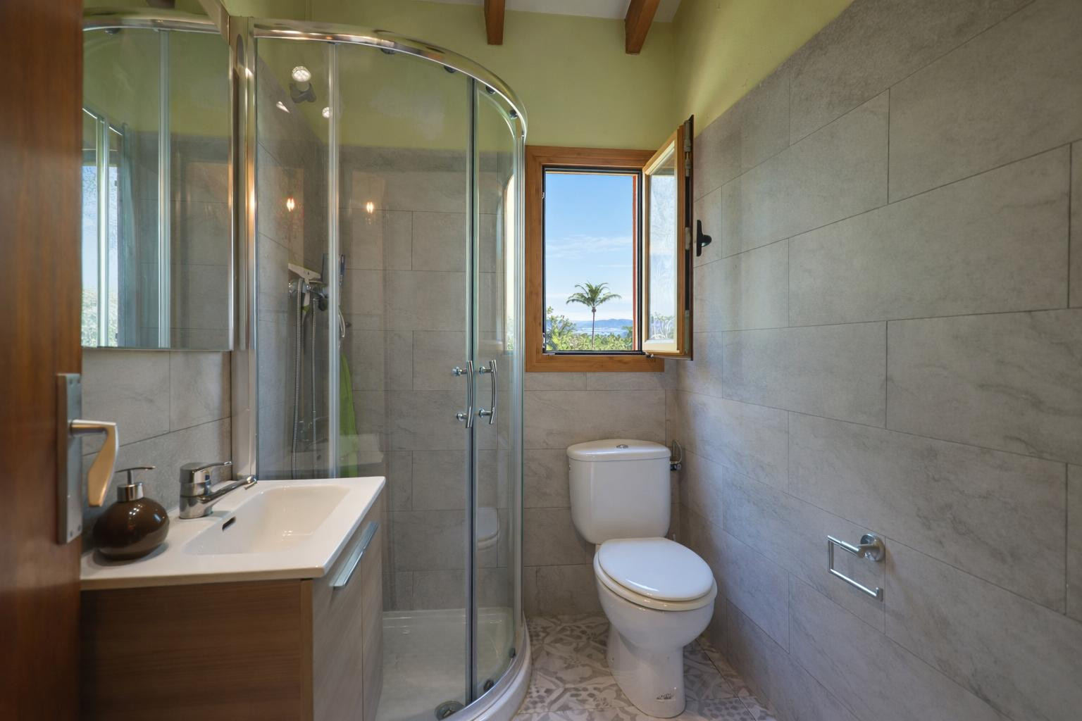 Salle de bains fonctionnelle avec douche à cabine courbe, carrelage gris et poutres en bois. La fenêtre apporte une lumière naturelle et une vue sur l'extérieur.