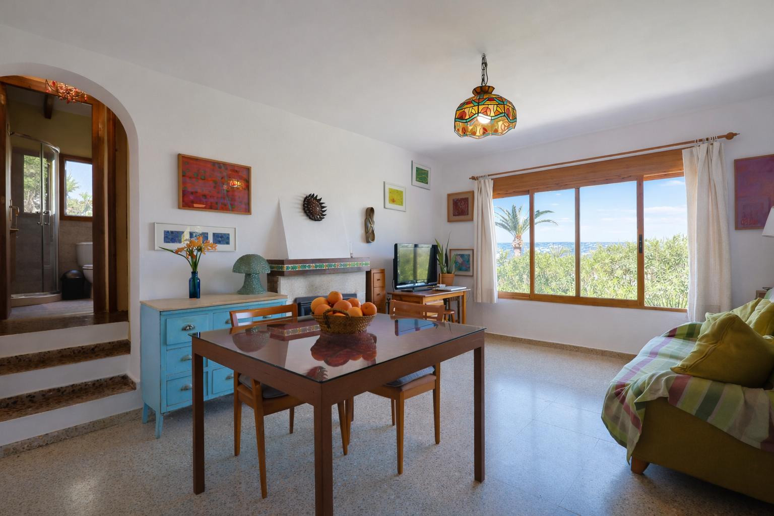 Salon-salle à manger avec cheminée, sol en terrazzo et vue sur mer. Une ouverture en arche mène à une salle de bain surélevée dotée d'une cabine de douche.