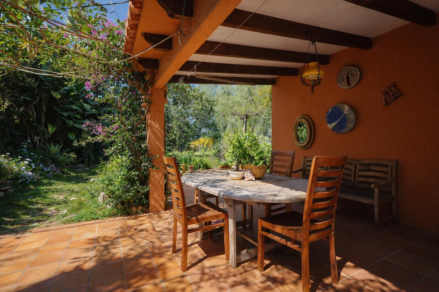 Terrasse couverte avec poutres apparentes, sol en terre cuite et coin repas extérieur offrant une vue sur le jardin méditerranéen verdoyant.