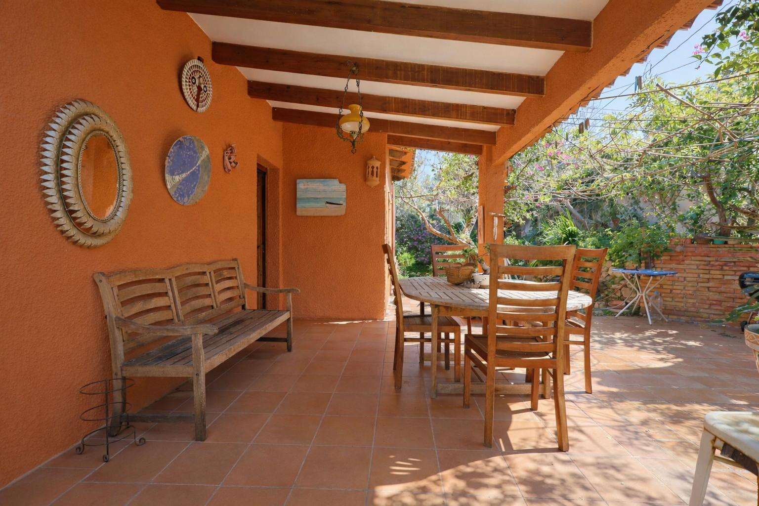 Terrasse couverte avec poutres en bois apparentes, sol en terre cuite et mobilier de jardin en bois dans un cadre méditerranéen.